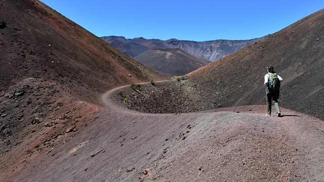 A person hikes through a red and brown trail through large hills.