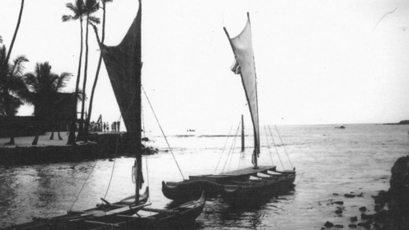 black and white image of two double-hulled canoes sit in a bay