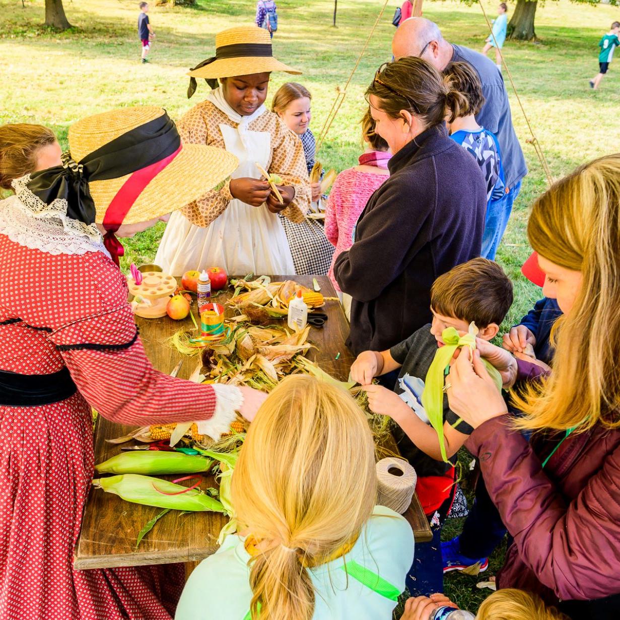 A living history intern helping visitors make corn husk dolls.