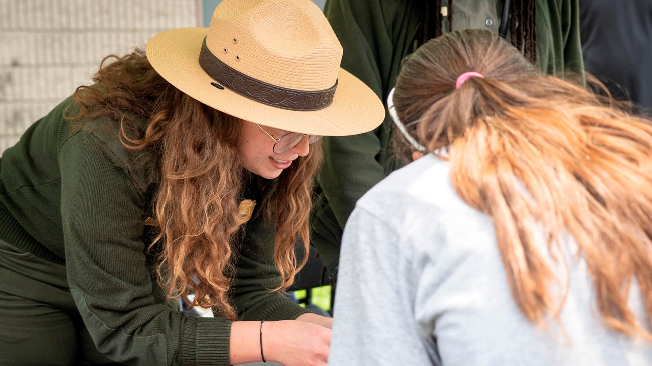A woman in a ranger uniform leans over a table and speaks to another woman. 