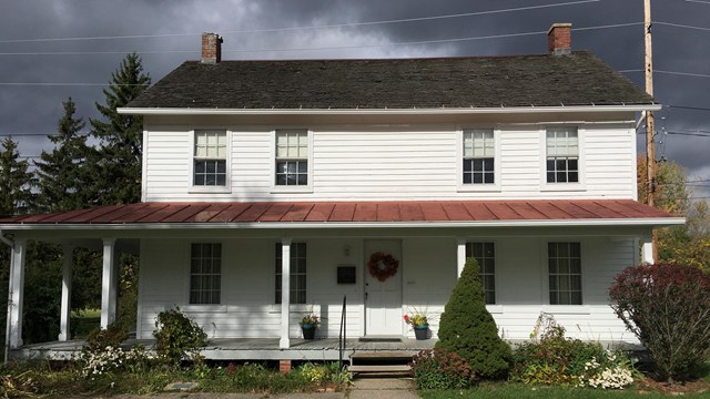 White, two-story house with thin columns on the front porch with shrubbery, and one red roof.