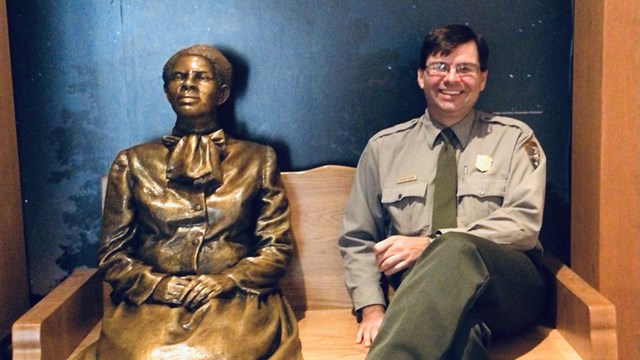 A park ranger sits beside a bronze statue of Harriet Tubman with a Tubman quote above them.