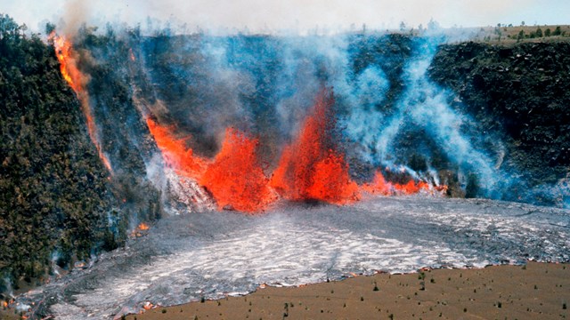 An erupting volcanic fissure at the base of a pit crater fountains orange molten lava and pools it