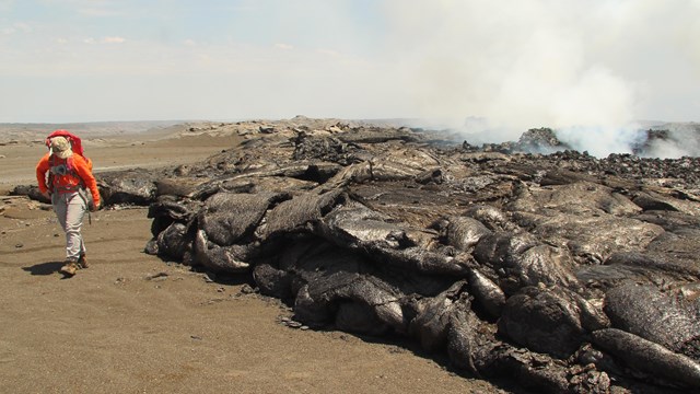 Figure wearing orange safety shirt walking next to dark smoking lava rock.