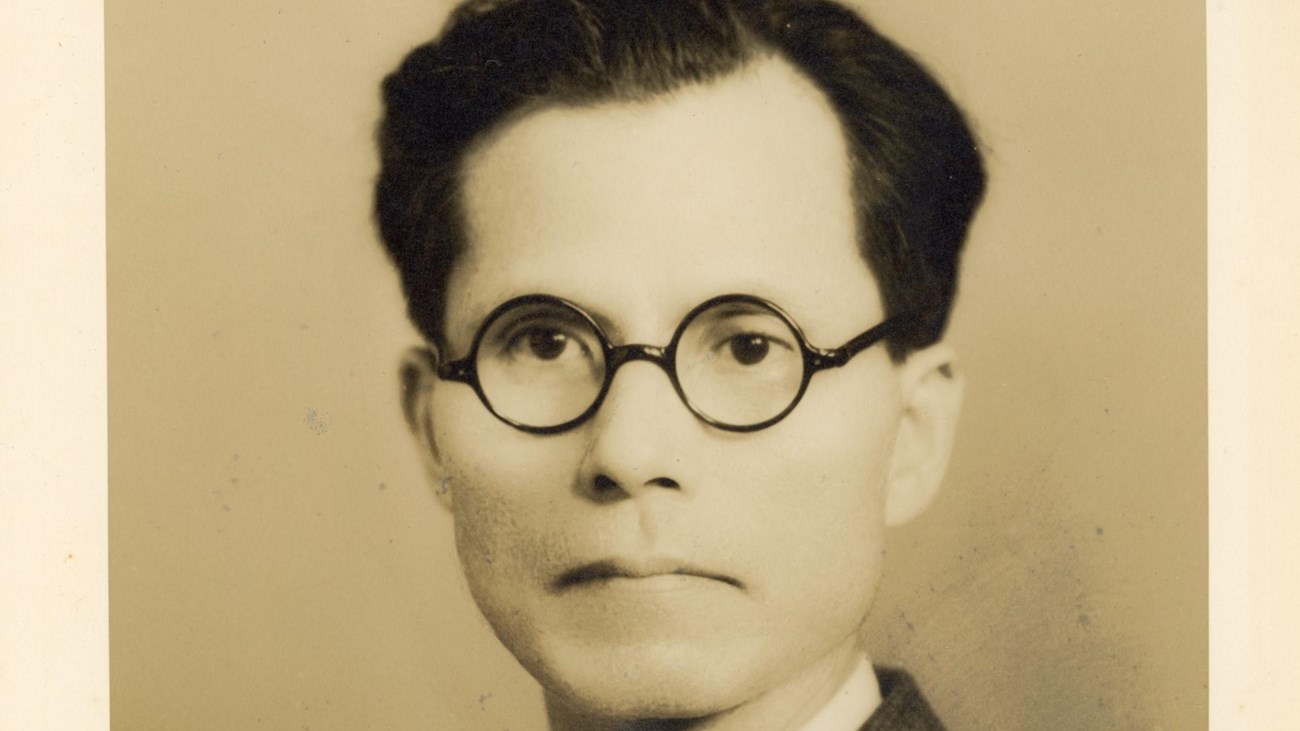 A black and white photograph of a Japanese man wearing glasses and a suit.