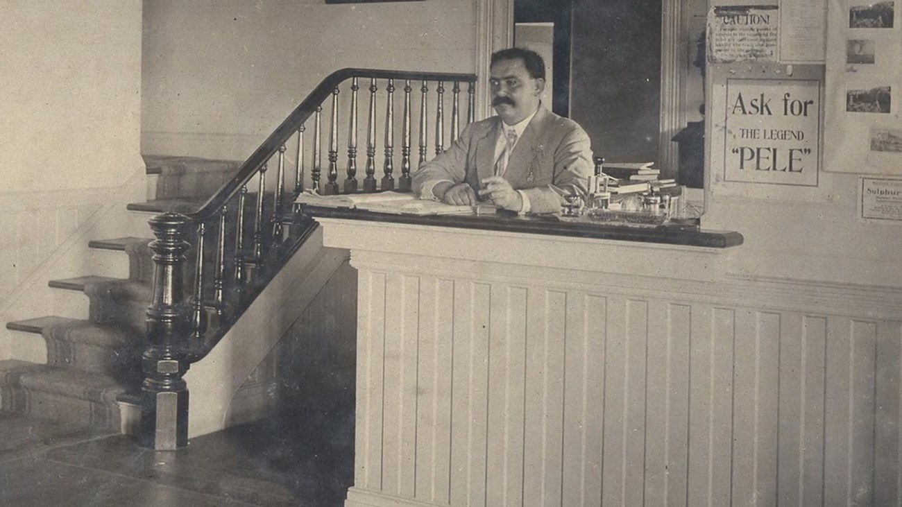 A man with mustache standing behind a counter next to a staircase and asign that says "Ask for Pele"