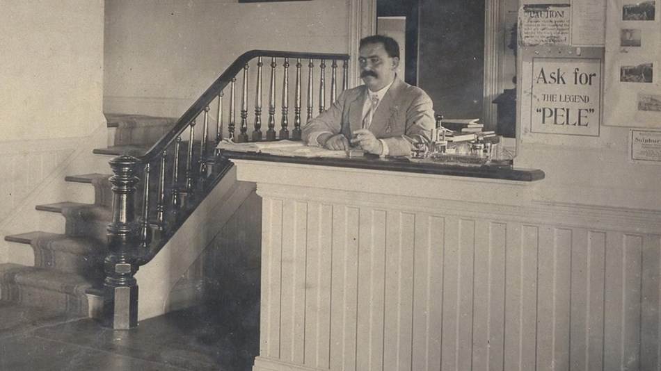 A man with mustache standing behind a counter next to a staircase and asign that says "Ask for Pele"