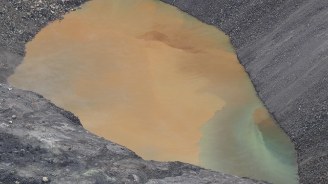 A brown lake in a volcanic crater with a swirl of turquoise on the lower half