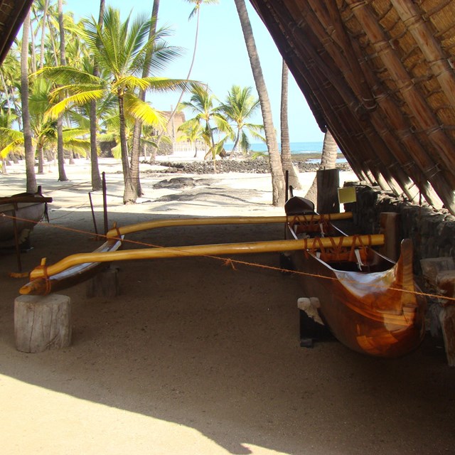 a hawaiian canoe sits under A-frame roof with palm trees and beach beyond