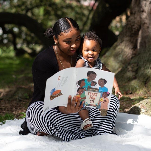 mother reads to baby while sitting on a blanket under a tree