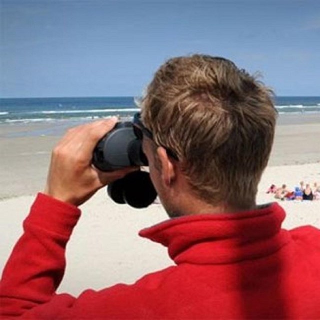One male life guard looking through binoculars and the other male lifeguard holding radio