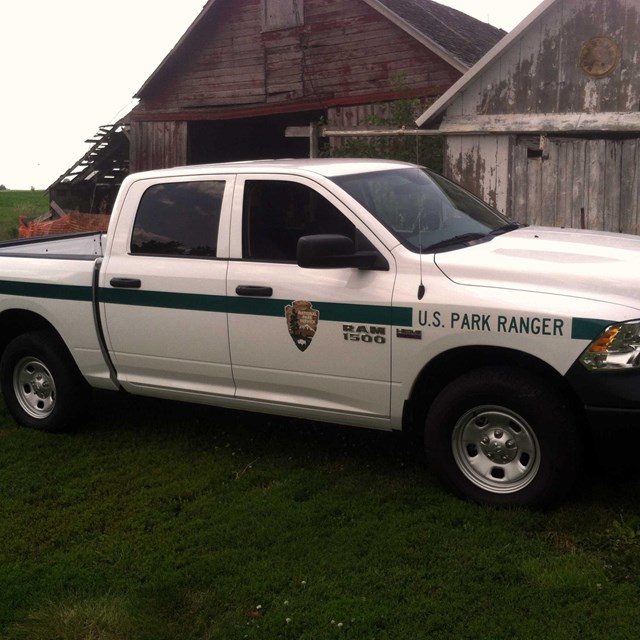 A park ranger patrol truck, painted white with a green stripe, is parked by a barn.