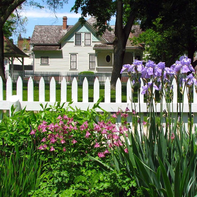A white picket fence separates purple flowers from a white house.