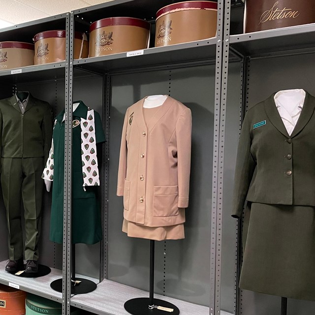 4 historical women's NPS uniforms display on a metal shelf with their round Stetson cases. 