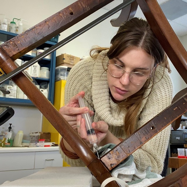 A woman pushes a syringe into the frame of a wooden chair that is propped up.