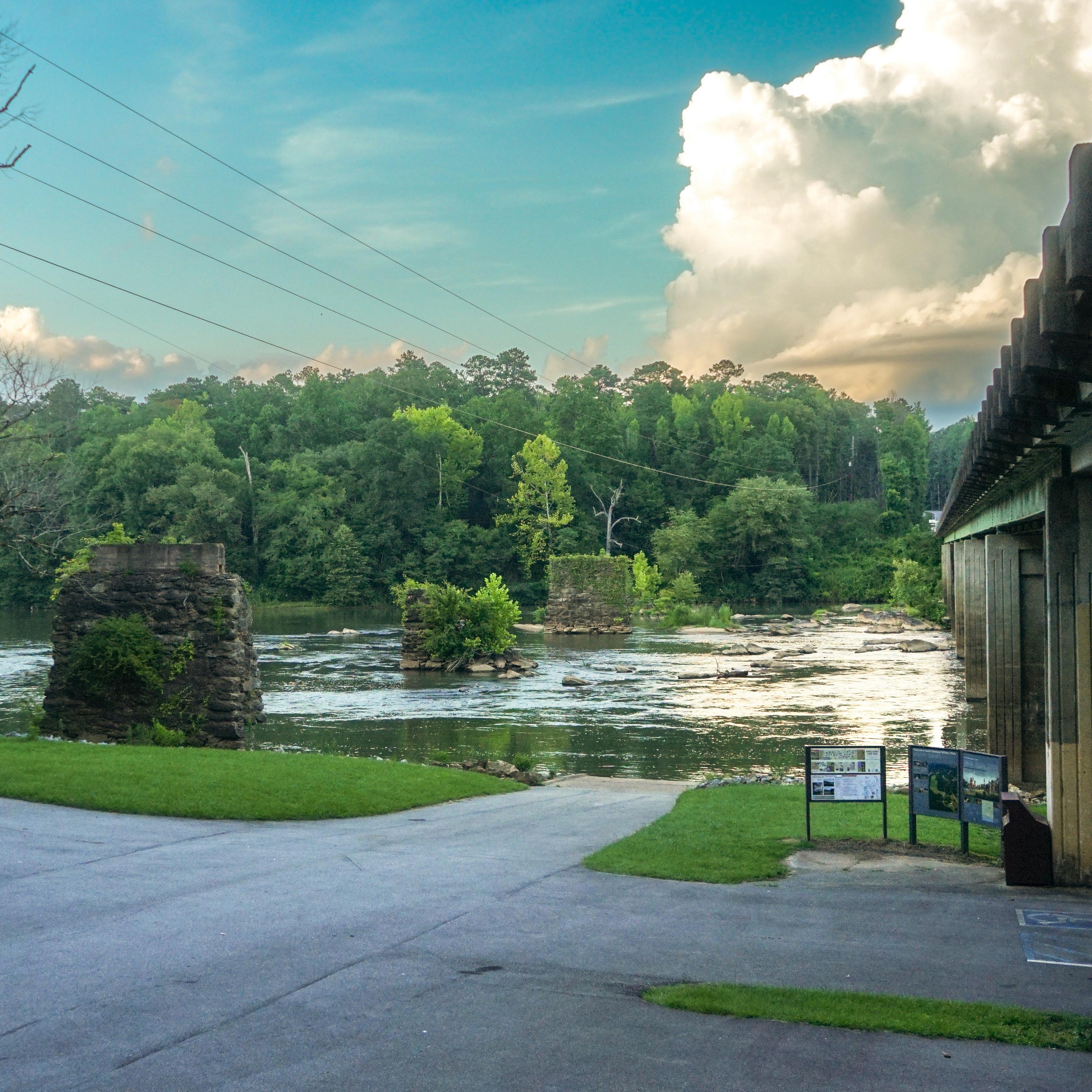 The image shows a small boat launch area next to a small parking lot, a large bridge, and a river.
