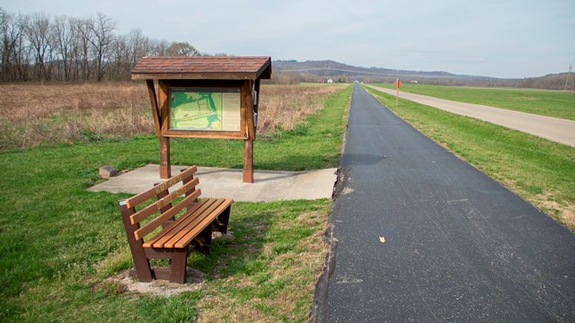 A long blacktopped path with a sign kiosk and bench to the left of it