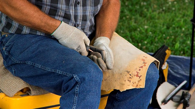 A person wearing blue jeans, a flannel shirt and gloves carves a piece of stone with a tool