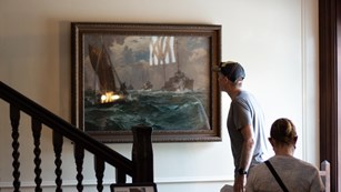 A man standing on a stair landing is looking closely at a naval painting.