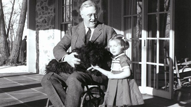 FDR seated in a wheelchair on the porch at Top Cottage. A young girl stands next to him.