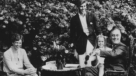 Four people of all ages seated and standing around a table on a lawn.