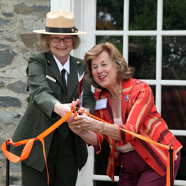 A park ranger in uniform and a woman jointly hold a pair of scissors cutting a ribbon.