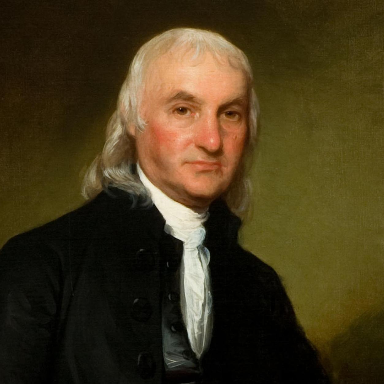 A seated man with long white hair wearing a dark suit and holding papers.