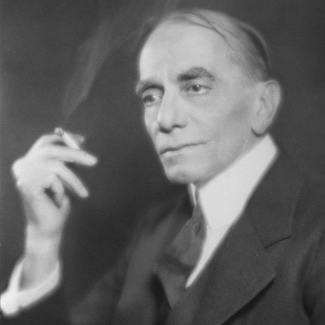 A photograph of a man in dark suit and tie holding a cigarette.