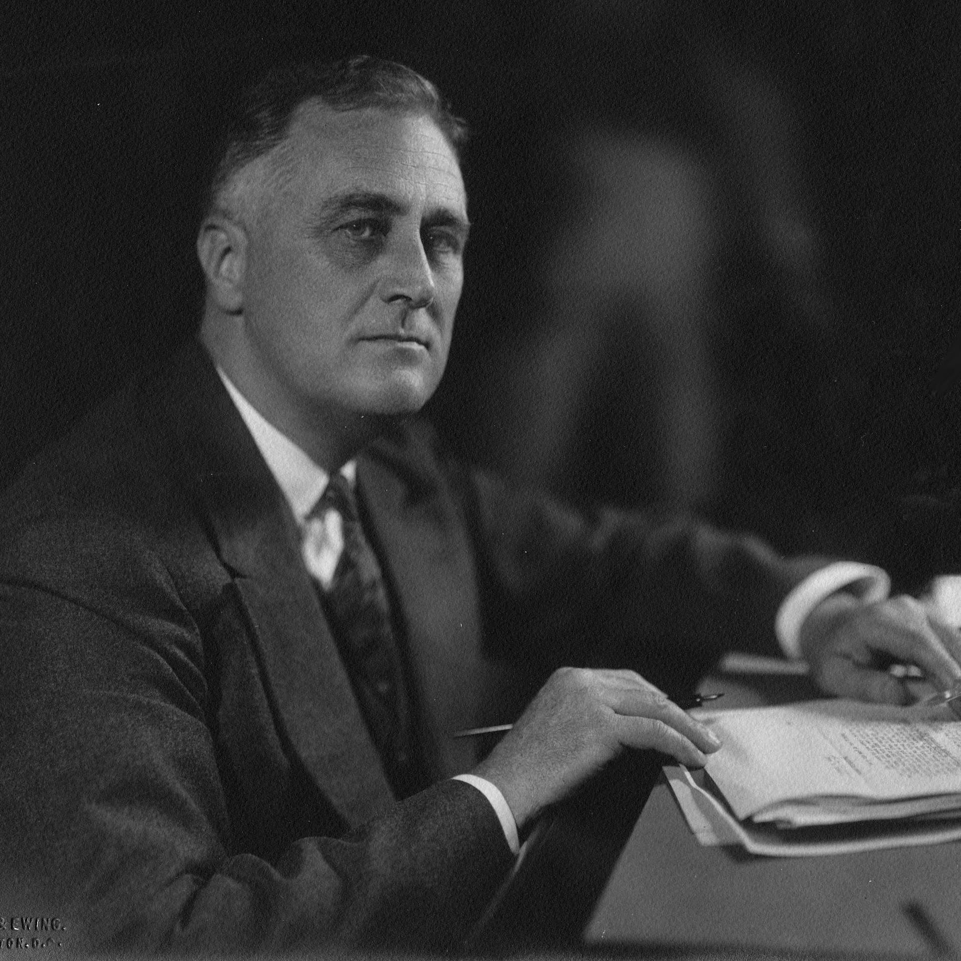 A man in a dark suit seated behind a desk with papers.