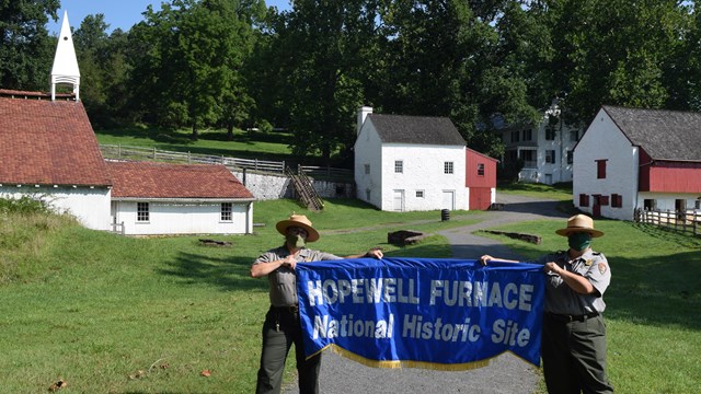 Two park rangers hold a Hopewell Furnace banner in the historic village.