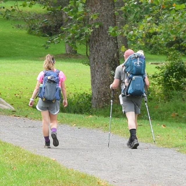 Two people with backpacks and hiking poles walk a dirt path 