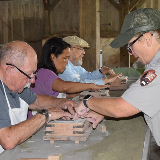 Park ranger working with a group of people. The peopel sit at a work bench and have boxes of sand.  