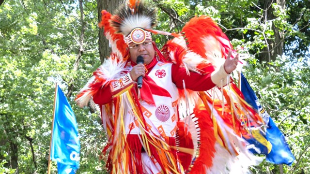 Man in traditional Native American dress speaks into microphone.