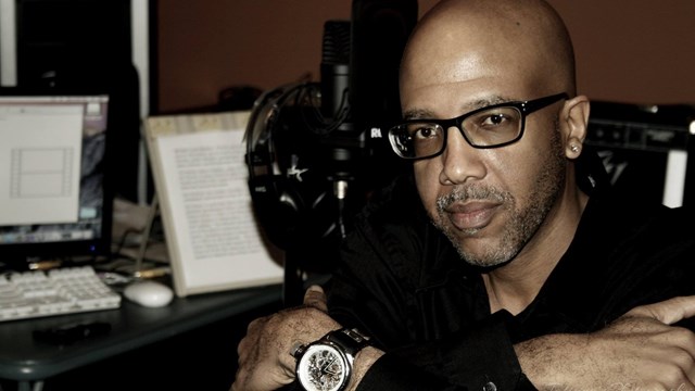 Black man looks into camera with crossed arms. Behind him is a computer and a text document.