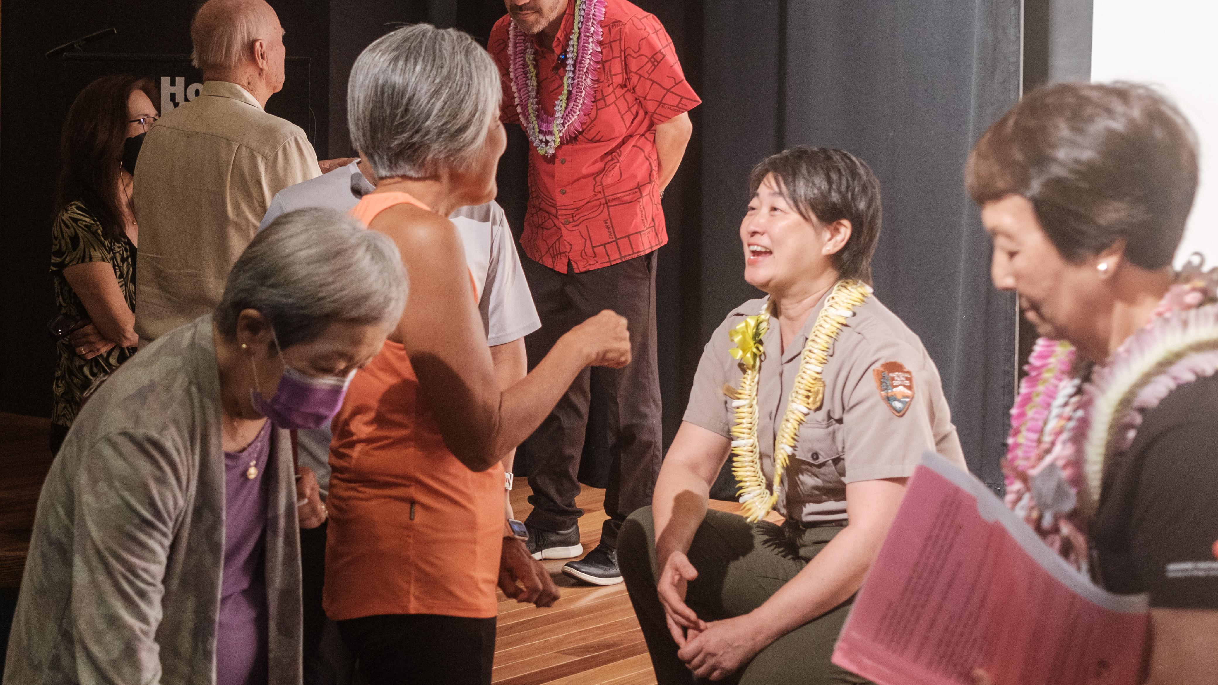 A park ranger in lei is crouching on a stage, smiling while talking to a woman standing