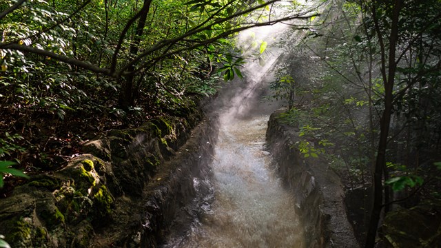 Thermal water flows down a concrete chute and is surrounded by green, lush trees.