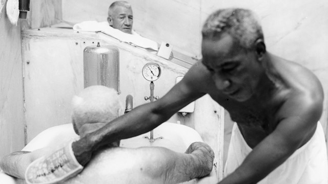 An African American man stands over a white man in a bathtub and rubs his back.