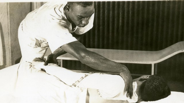 An African American man stands over a white man in a towel. He is rubbing the white man's shoulder.