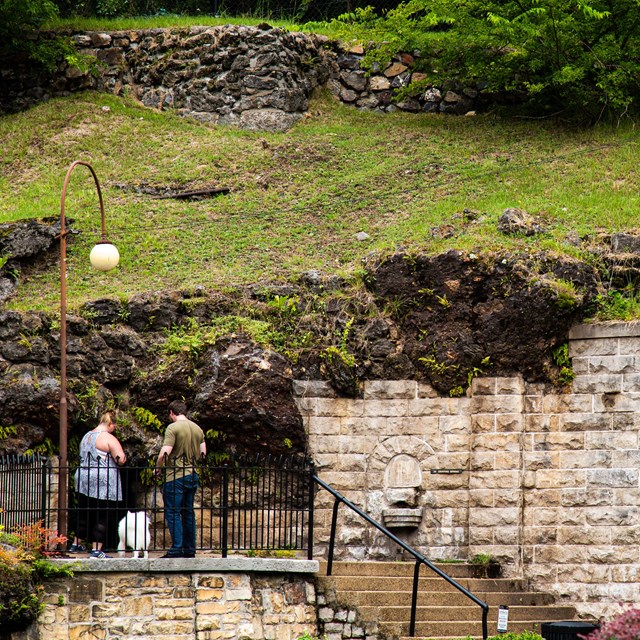 A couple stands on top of a few steps looking down into a cavern in the hillside.