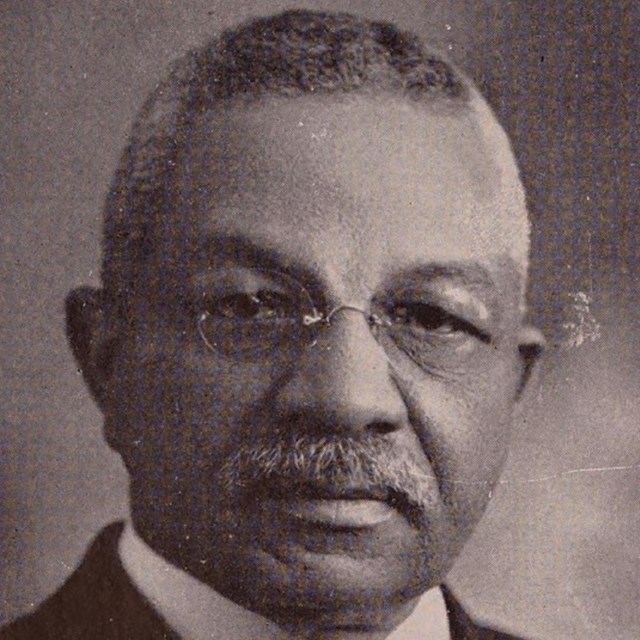 African American man wears a white shirt, dark tie, black jacket, and glasses.