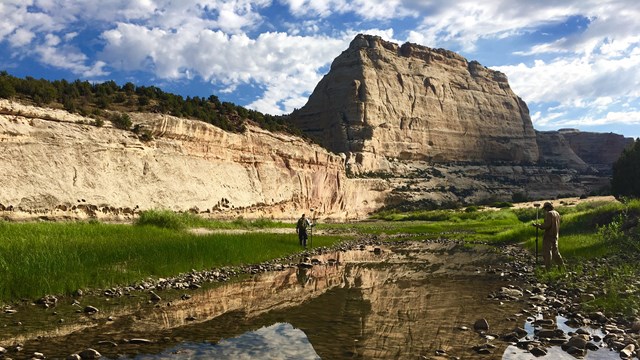 Two people stand along a river that reflects a rock monolith