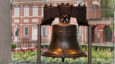 Color photo of the Liberty Bell with Independence Hall visible through the glass behind it.
