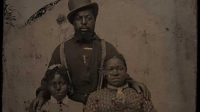 A woman and girl sitting and a man standing behind them, all facing towards viewer