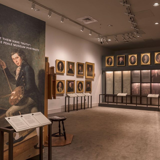 Color photo of exhibit area with a picture of one man (foreground) with rows of portraits behind.