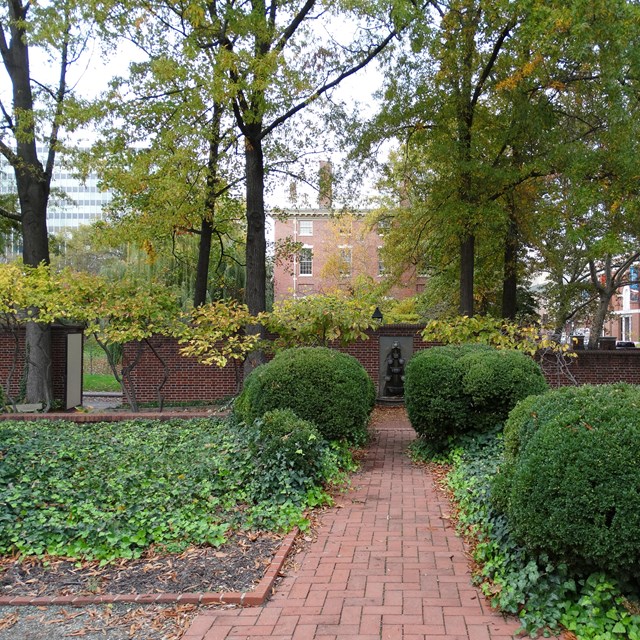 Bushes and leafy foliage line a central brick path leading to a wall mounted fountain.