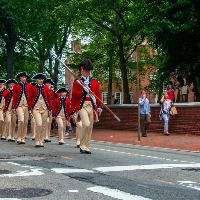 Color photo of a group of men marching in a line dressed in British military uniforms.