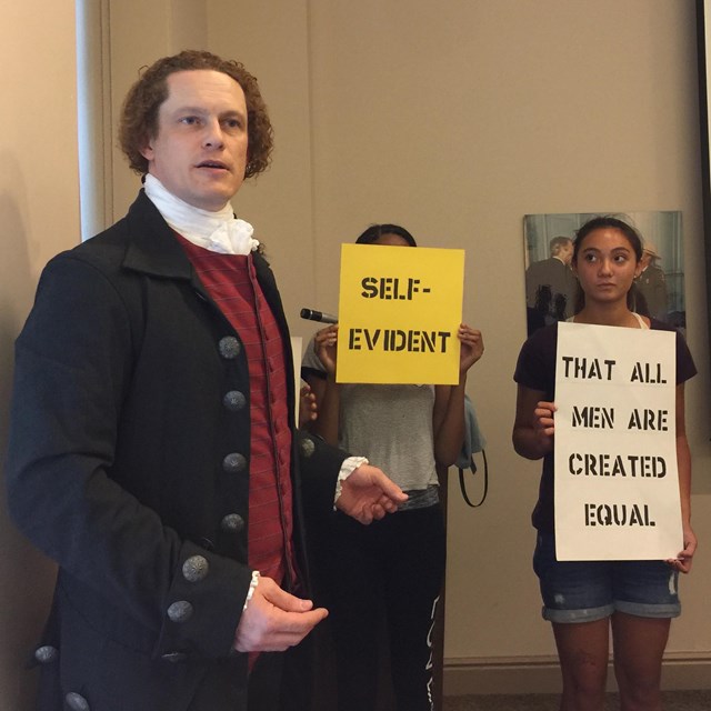 Color photo of a man in colonial costume in front of teens holding phrases from the Declaration.