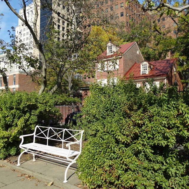 Color photo of white bench among foliage with a red brick house in the background.