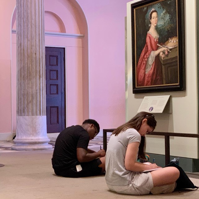 Color photo of three students seated on the floor below a portrait of a woman from the 1700s.