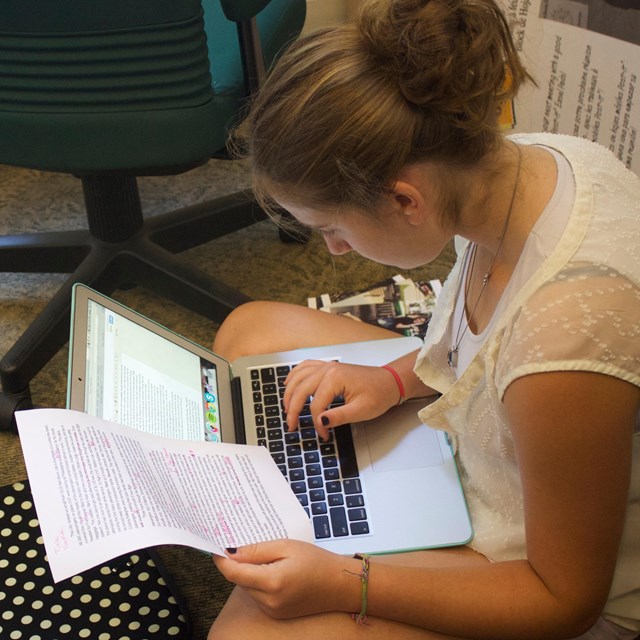 Color photo of a teen girl sitting on the floor with a laptop computer on her lap.